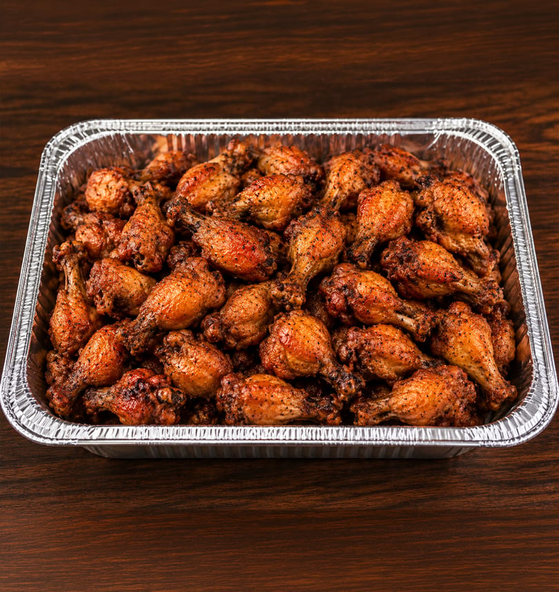 A large foil pan filled with numerous golden-brown, seasoned chicken drumettes on a dark wooden surface.