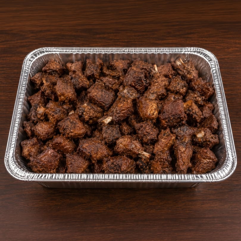 A foil pan filled with many pieces of dark brown, saucy bone-in meat, on a dark wooden surface.
