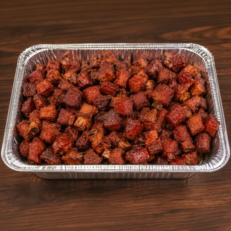 Close-up of a foil pan filled with glistening, dark red barbecue burnt ends on a wooden table.